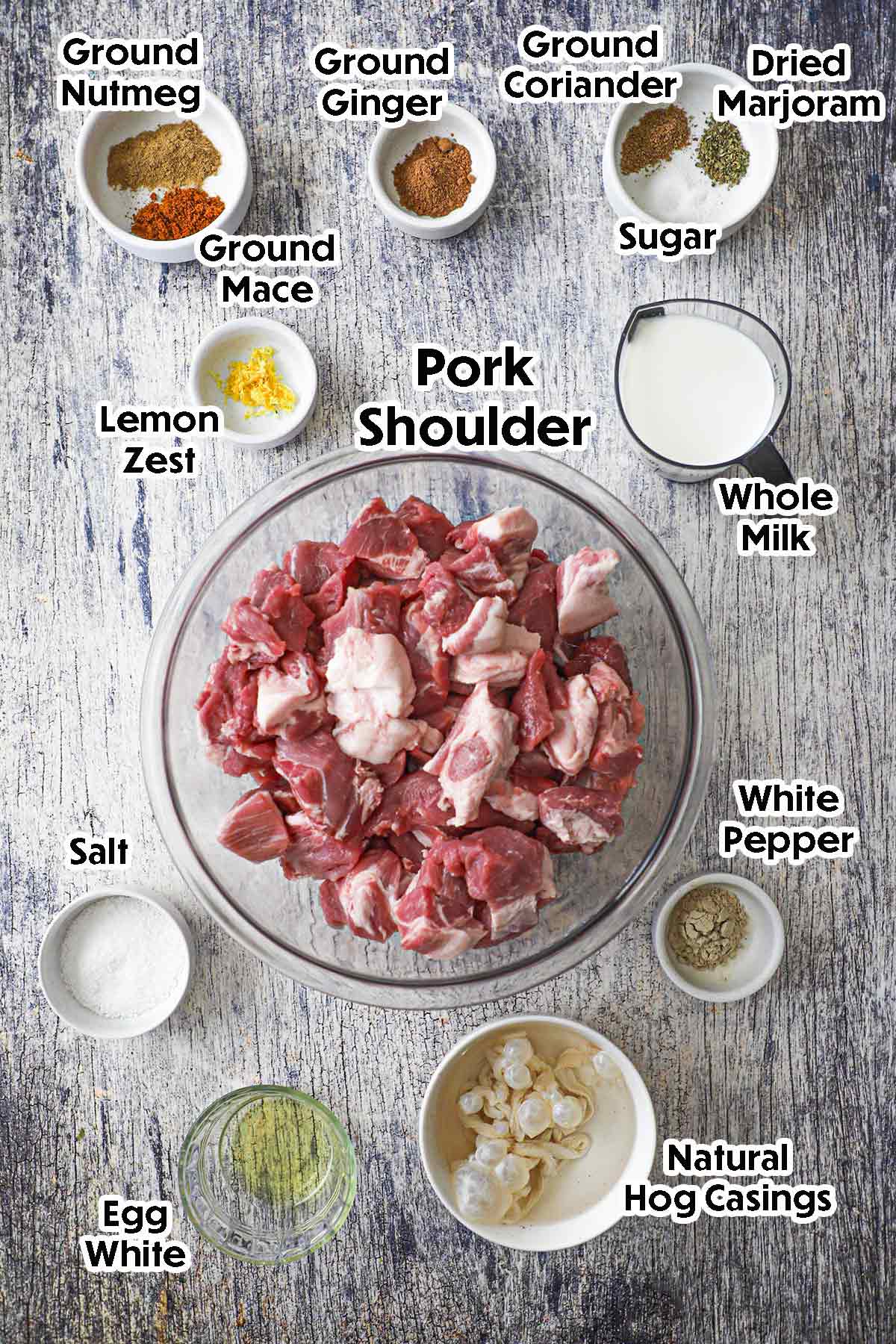 An arrangement of ingredients for homemade bratwurst on a grey-wooden background including pork shoulder, milk, lemon zest, hog casings, an egg white, spices, and seasonings.