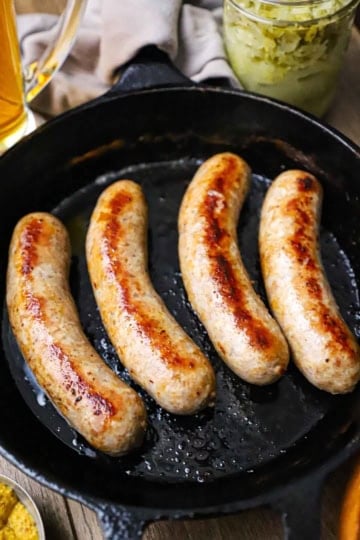 A straight-on view of four links of seared homemade bratwurst in a black cast-iron skillet with a glass of beer and a jar of sauerkraut sitting nearby.