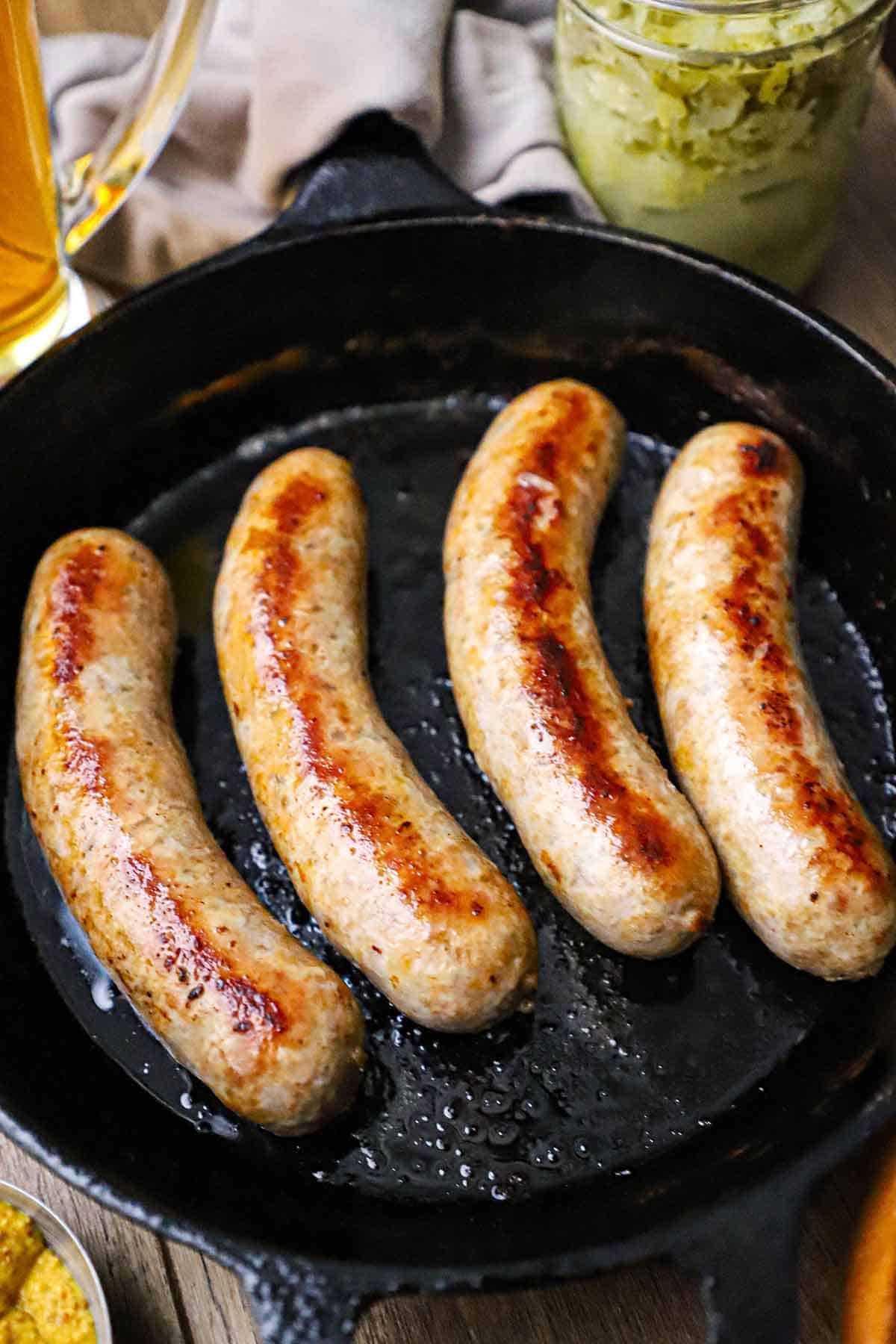 A straight-on view of four links of seared homemade bratwurst in a black cast-iron skillet with a glass of beer and a jar of sauerkraut sitting nearby.