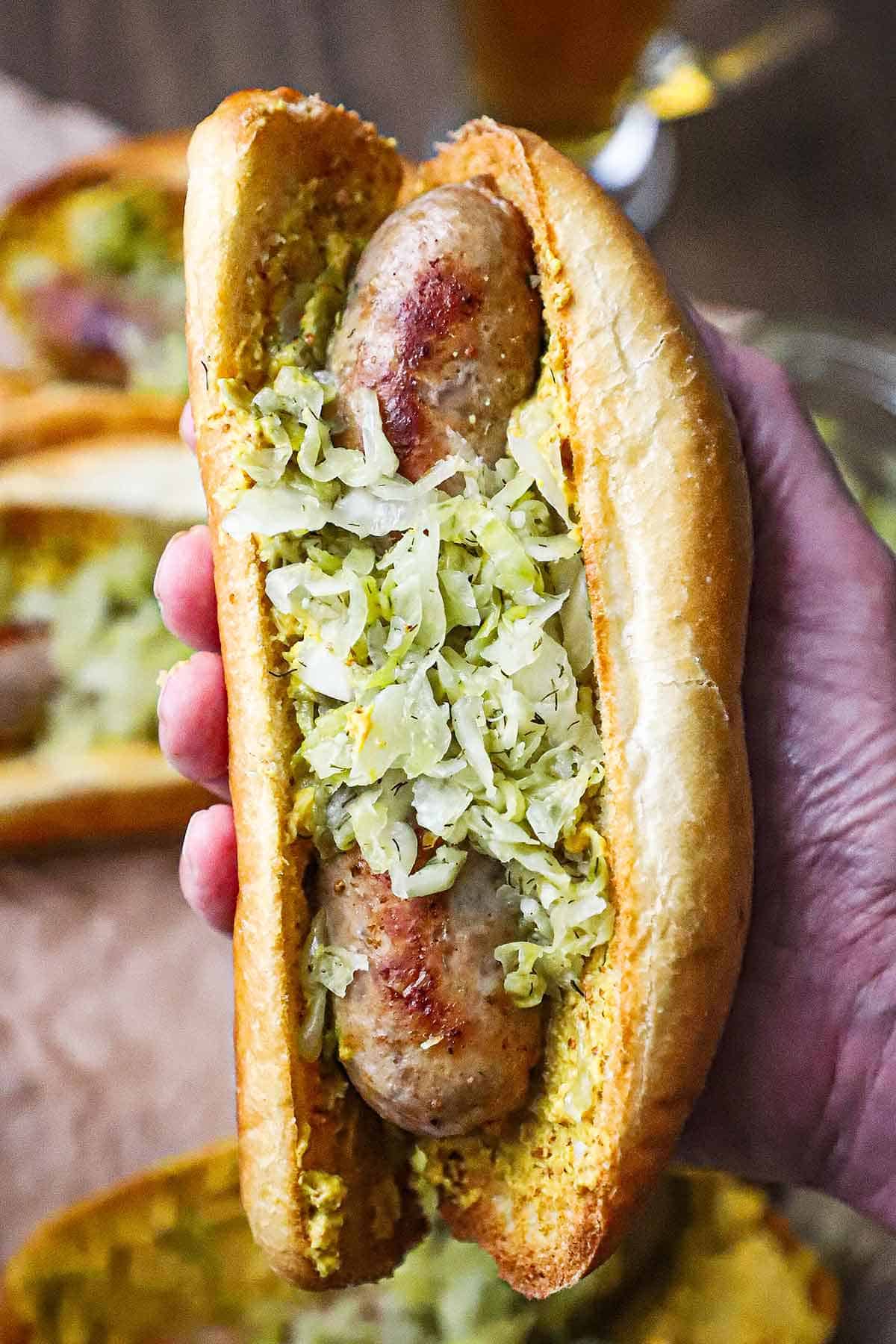 A person holding a homemade bratwurst sausage in a toasted roll with brown mustard and topped with homemade sauerkraut.