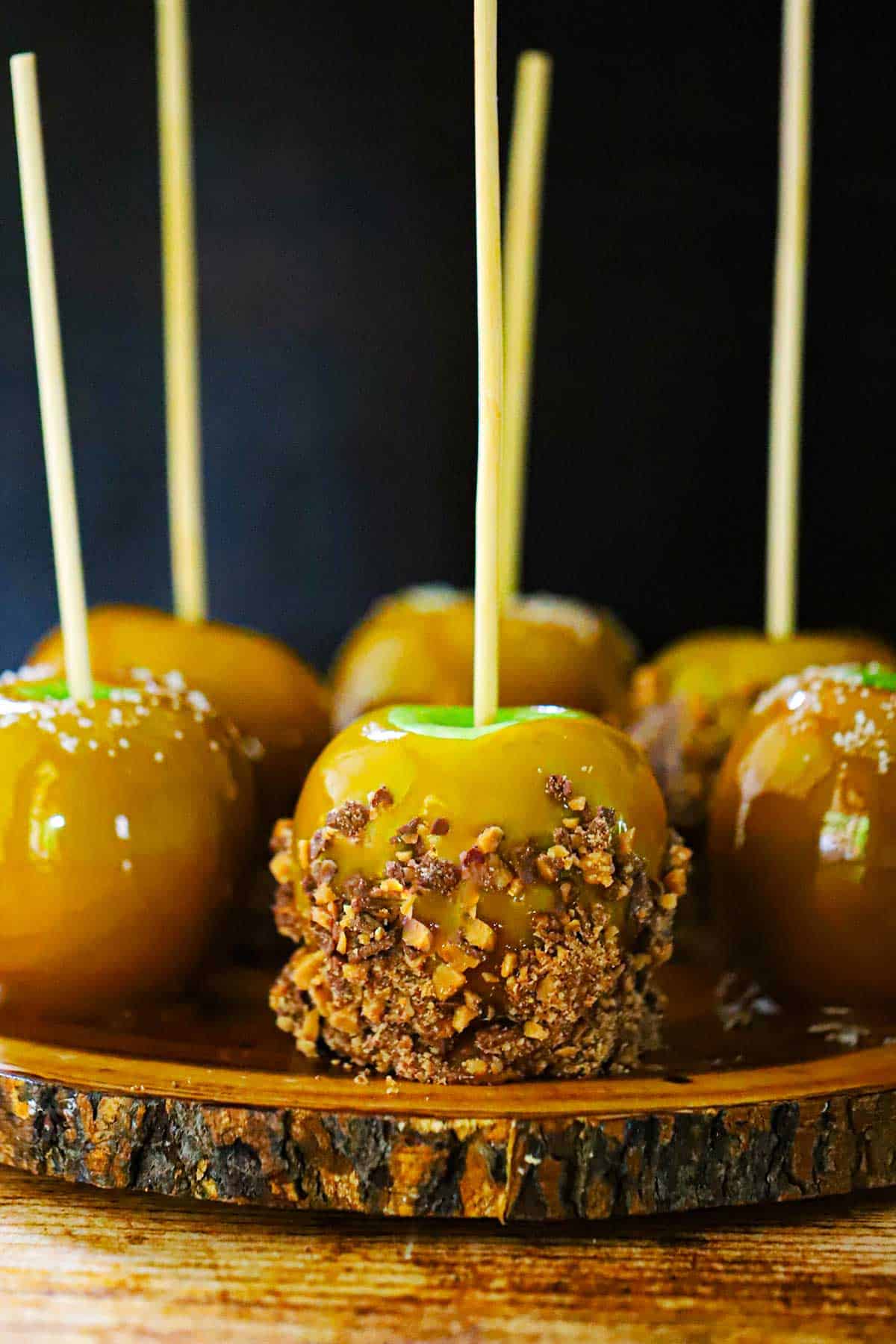 A straight-on view a a grouping of Honey Caramel Apples with Toffee Bits or Sea Salt sitting on a circular wooden slab all with wooden sticks inserted into the tops of each apple.