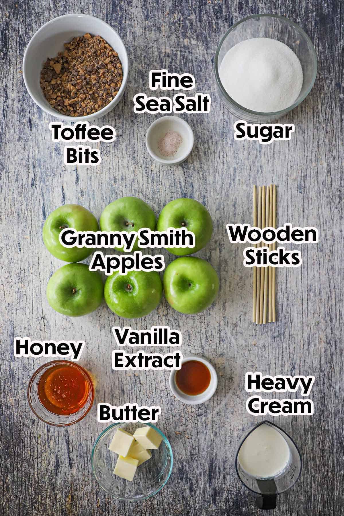 An arrangement of ingredients for Honey Caramel Apples on a grey-wooden background including Granny Smith apples, honey, sugar, salt, butter, cream, vanilla extract, toffee bits, and wooden sticks.