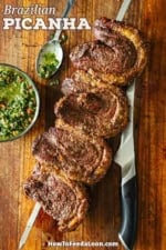 Four freshly cooked Brazilian Picanha steaks on a skewer resting on a cutting board with a bowl and spoon filled with homemade chimichurri sauce next to it.