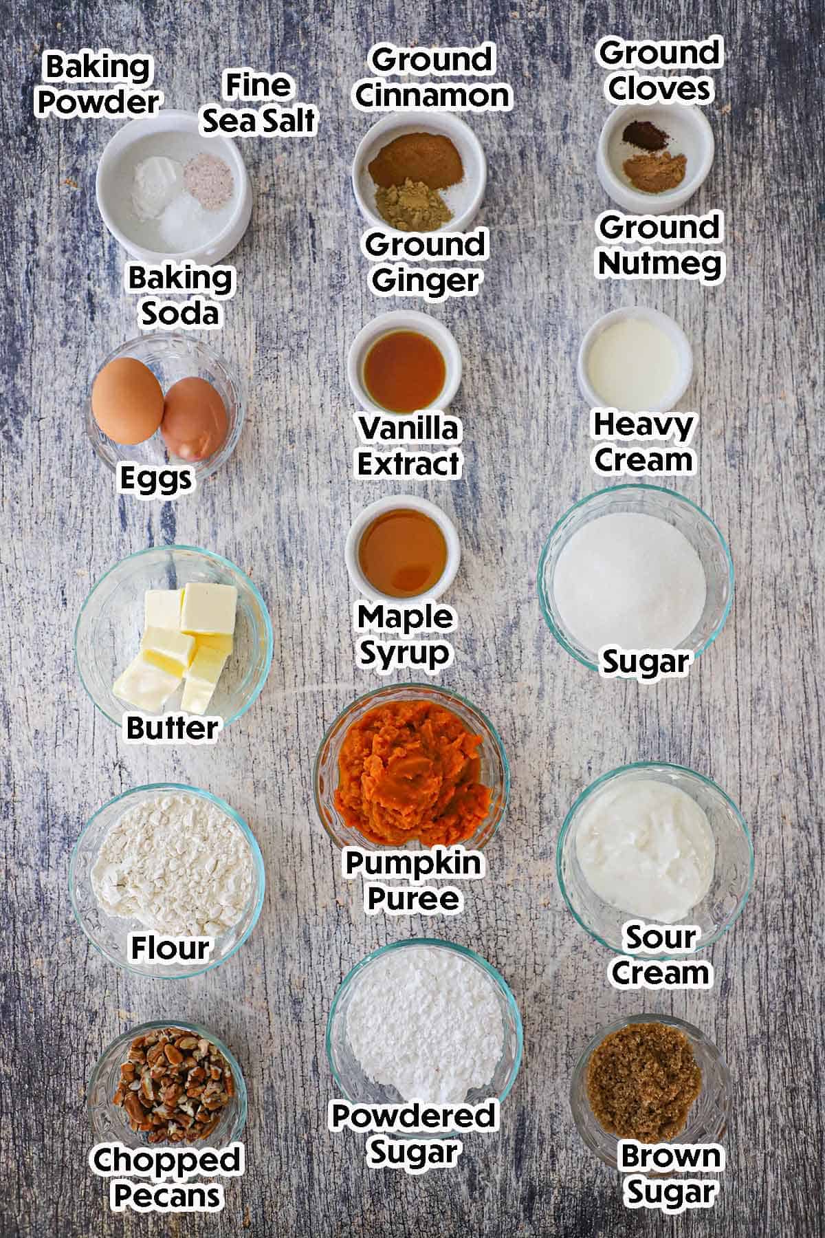 An arrangement of ingredients for Pumpkin Spice Coffee Cake on a grey-wooden background including flour, pumpkin purée, sour cream, butter, eggs, pecans, sugar, sour cream, and spices.