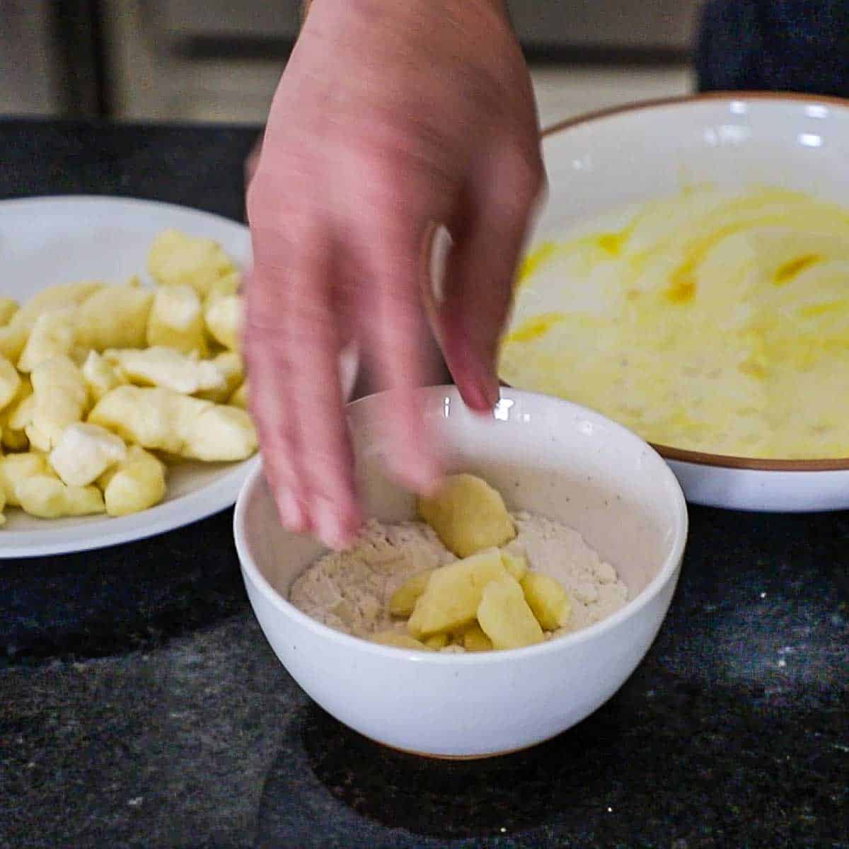 A person tossing cheese curds in a small bowl filled with flour next to a plate filled with more cheese curds and another shallow bowl filled with combined eggs and buttermilk.