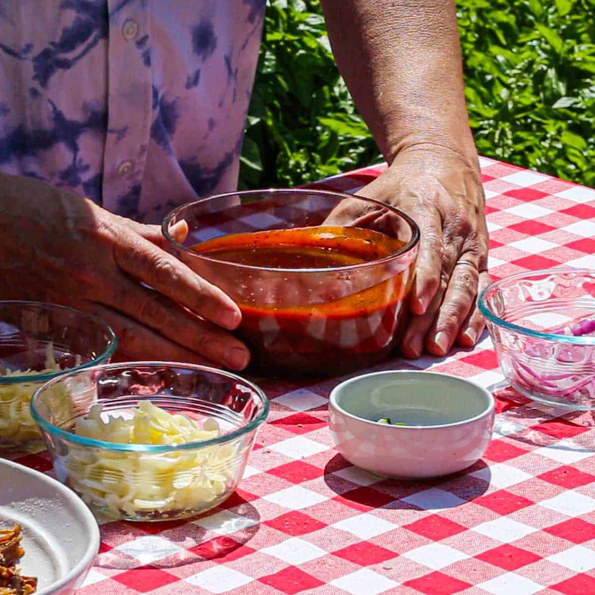 A person holding a glass bowl that is filled with homemade barbecue sauce on a table that is covered with a red-checkered table cloth.