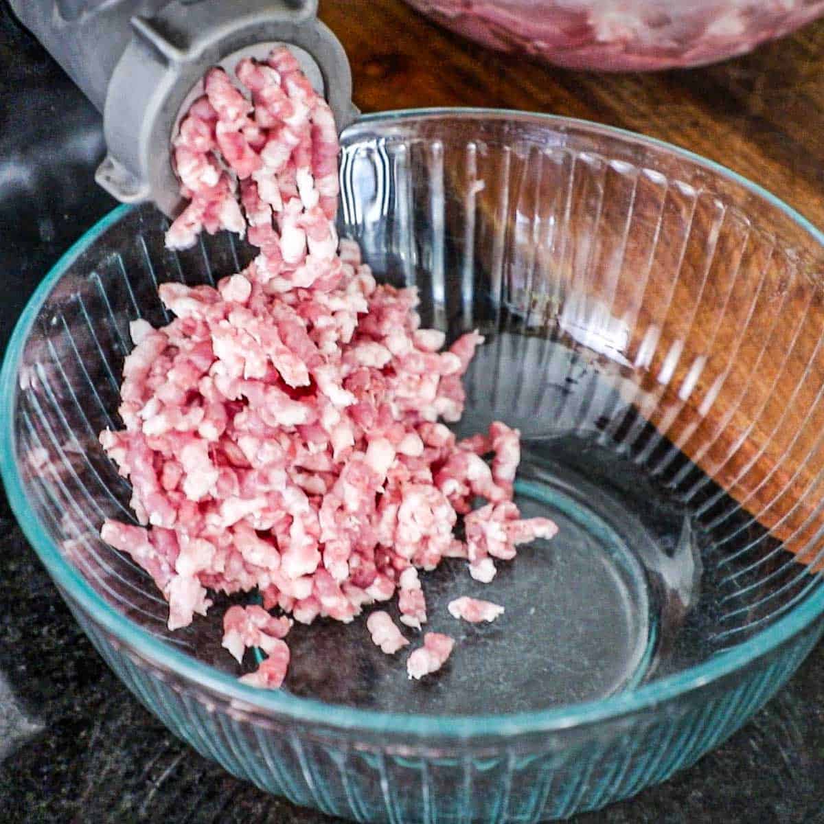 Ground pork being exuded from a meat grinder into a large glass bowl.