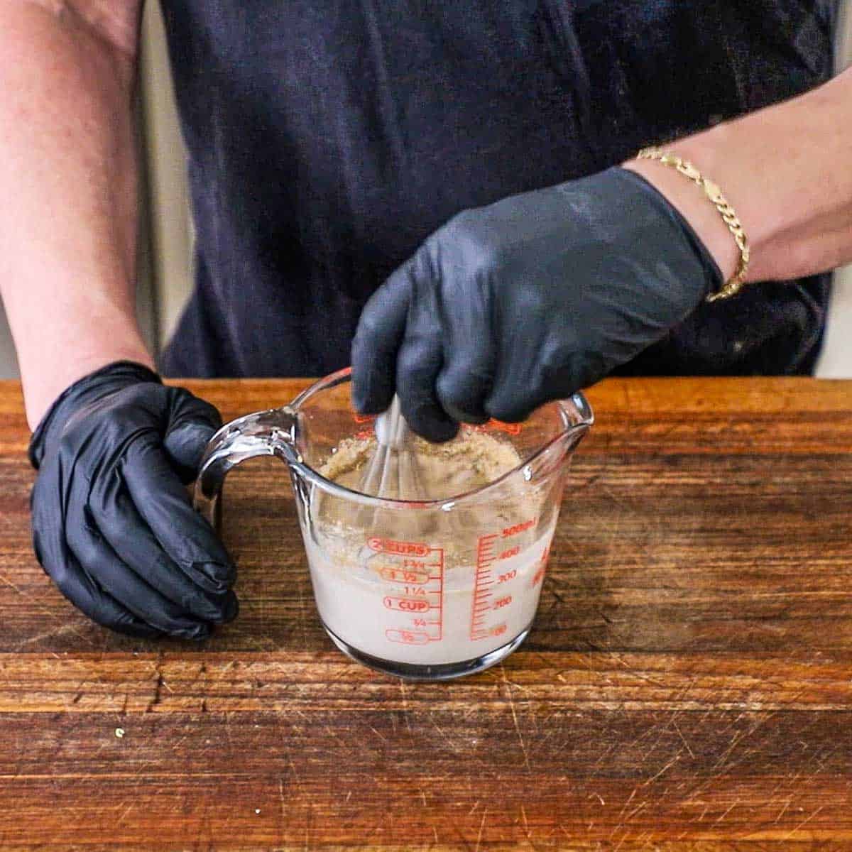 A person wearing black kitchen gloves using a small whisk to combine whole milk with bratwurst spices in a glass measuring cup.