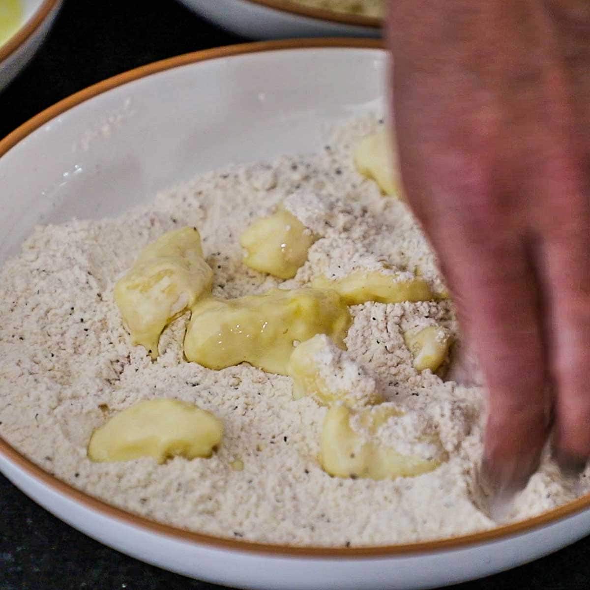 A person using his fingers to toss cheese curds that are coated with a flour and egg mixture in a shallow bowl that is filled with seasoned flour.