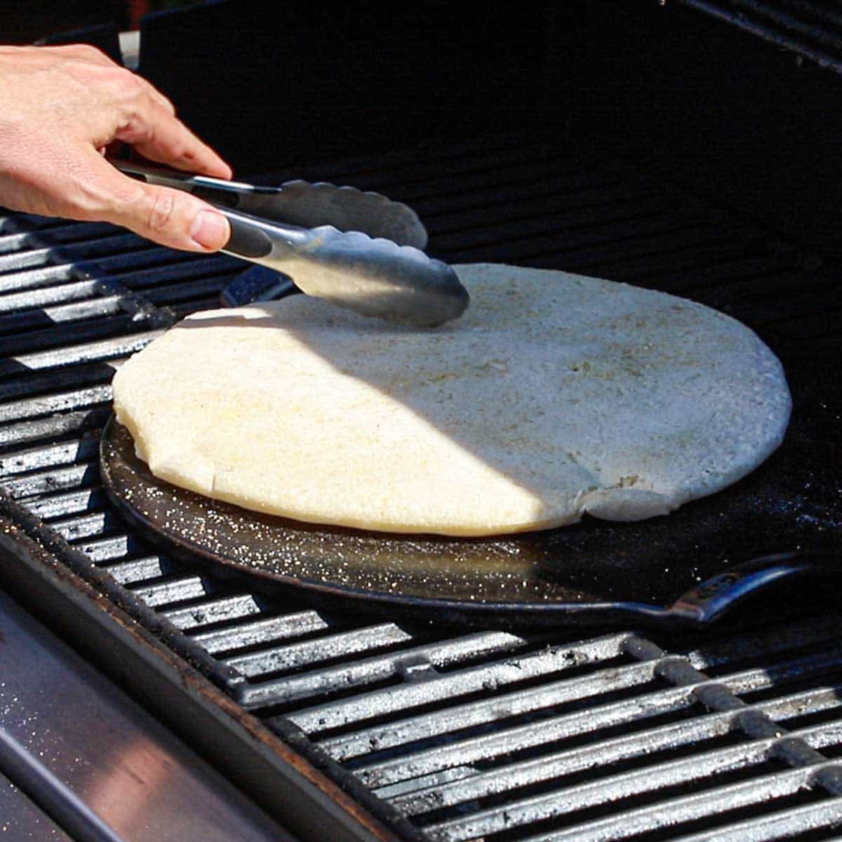 A person using a pair of tongs to flip a partially cooked round pizza dough on a hot pizza stone that is resting on the grates of a gas grill.
