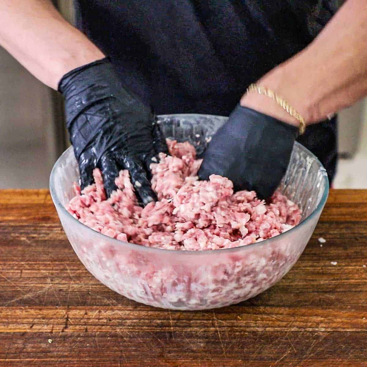 A person wearing black kitchen gloves using his hands to mix ground pork with whole milk that has been combined with bratwurst seasonings.