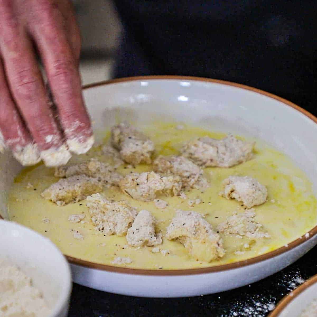 A person using his fingers to toss egg and flour battered cheese curds in a shallow bowl that is filled with an egg and buttermilk mixture.