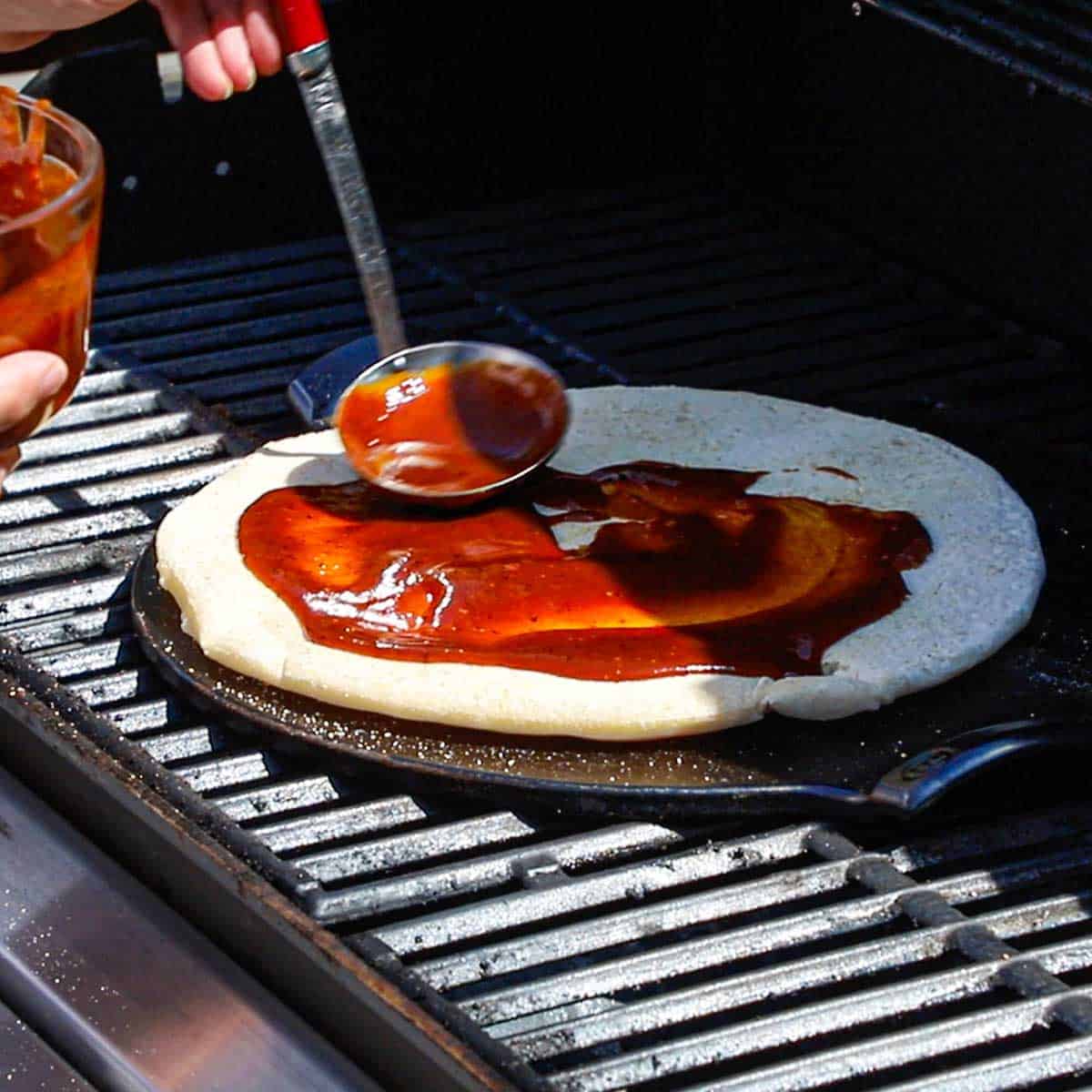 A person using a ladle to add homemade barbecue sauce over the surface of a partially cooked pizza dough that is on a hot pizza stone on the grates of a gas grill.