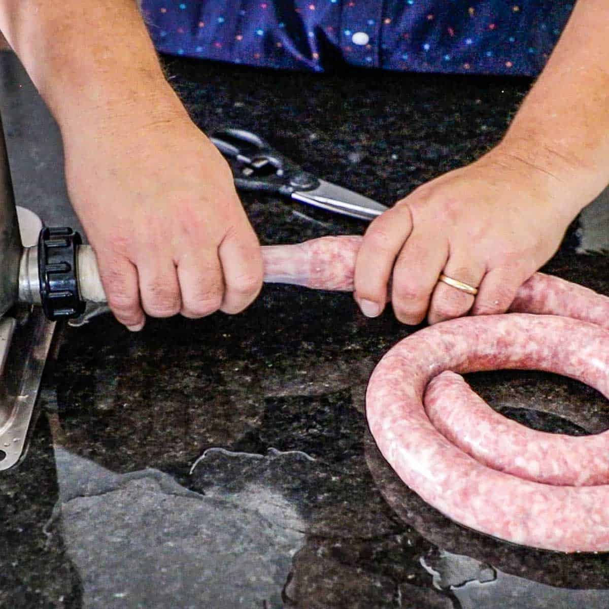 A person using his two hands to guide raw bratwurst filling into natural hog casings that are attached to a sausage stuffer.