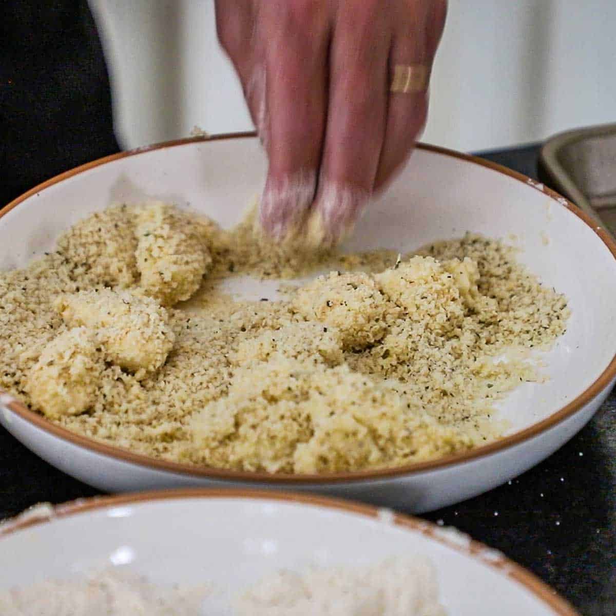 A person tossing battered cheese curds in a shallow bowl filled with Panko breadcrumbs mixed with dried oregano.