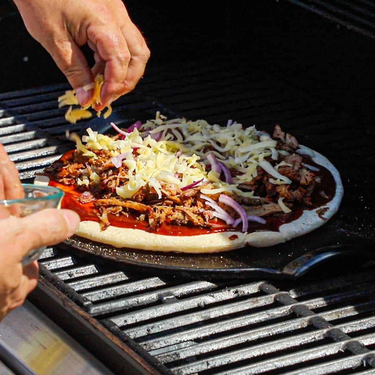 A person sprinkling shredded cheese over the top of a pulled pork pizza that is being cooked on a hot pizza stone on the grates of a gas grill.