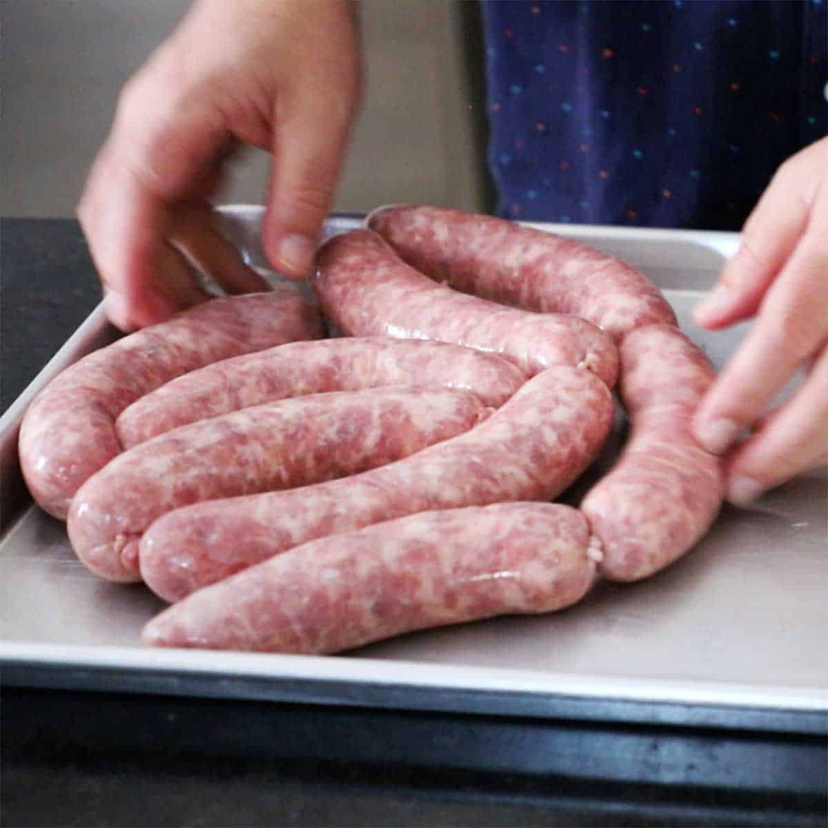 A person placing freshly stuffed raw homemade bratwursts onto a metal sheet pan.