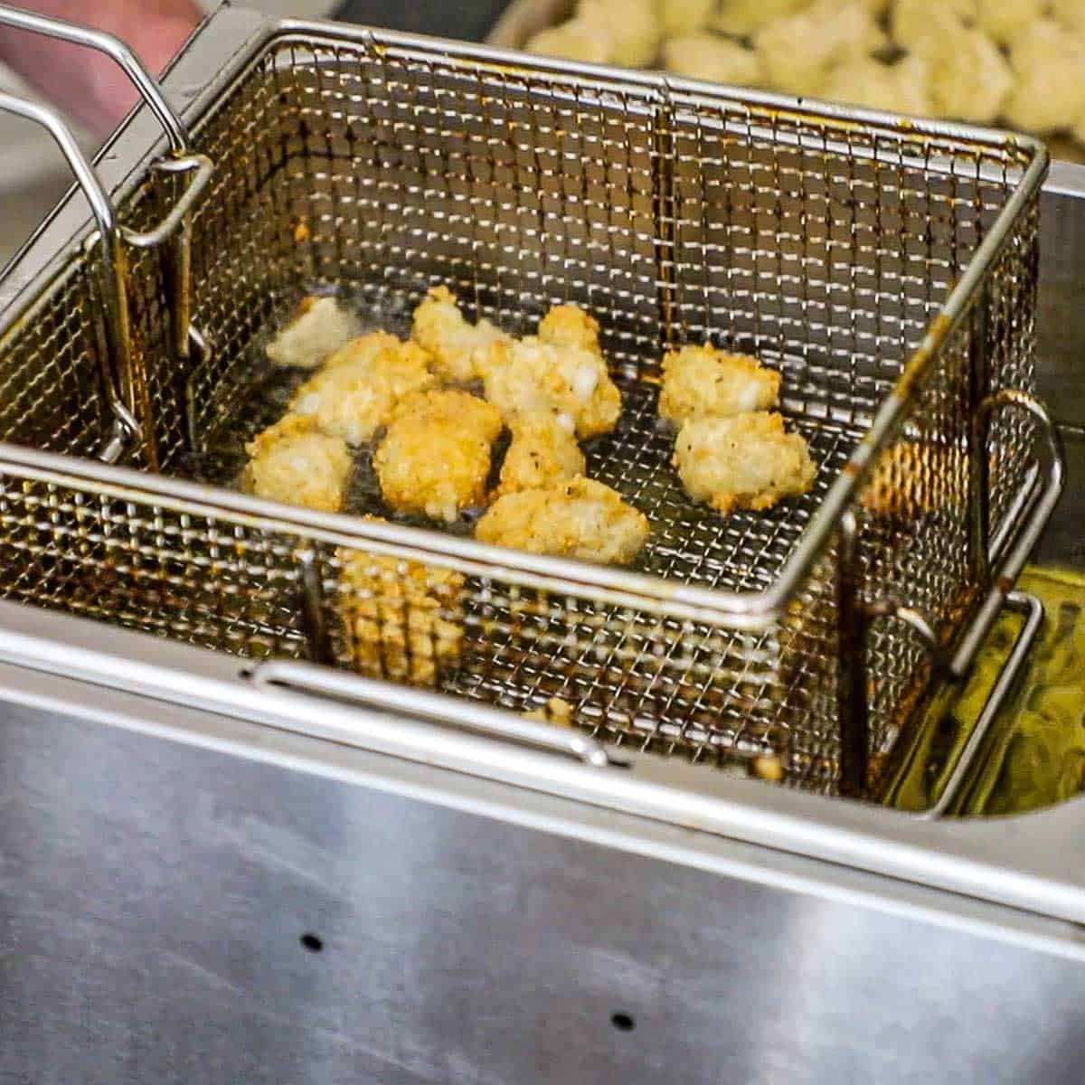 A person raising a fry basket filled with fried cheese curds from a deep-fryer.