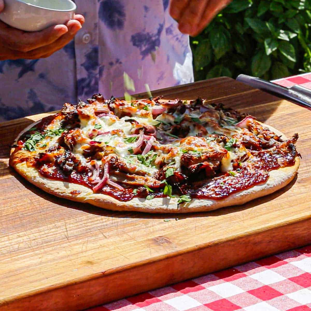 A person sprinkling chopped Italian parsley over the top of a freshly prepared Grilled Pulled Pork Pizza that is resting on a wooden cutting board.