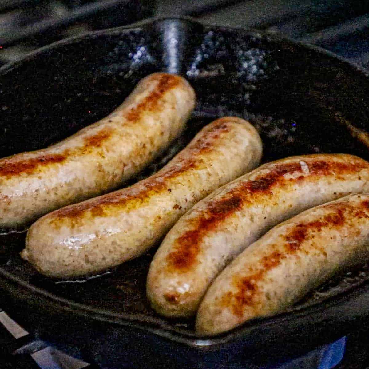 A close-up view of four links of homemade bratwurst that are being seared in a cast-iron skillet on a gas stove.