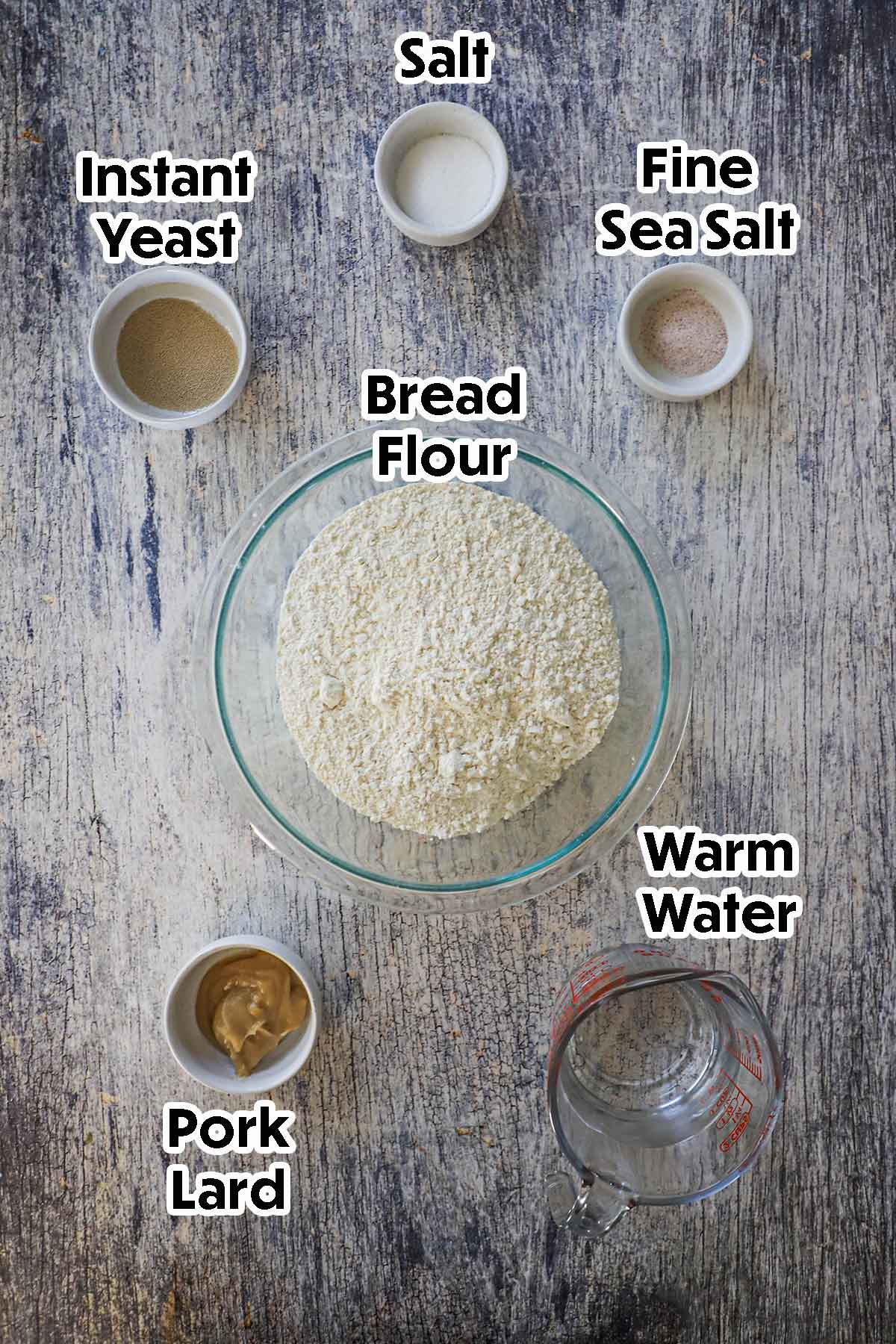 An arrangement of ingredients for homemade telera rolls on a grey wooden background including bread flour, yeast, pork lard, water, and sugar.