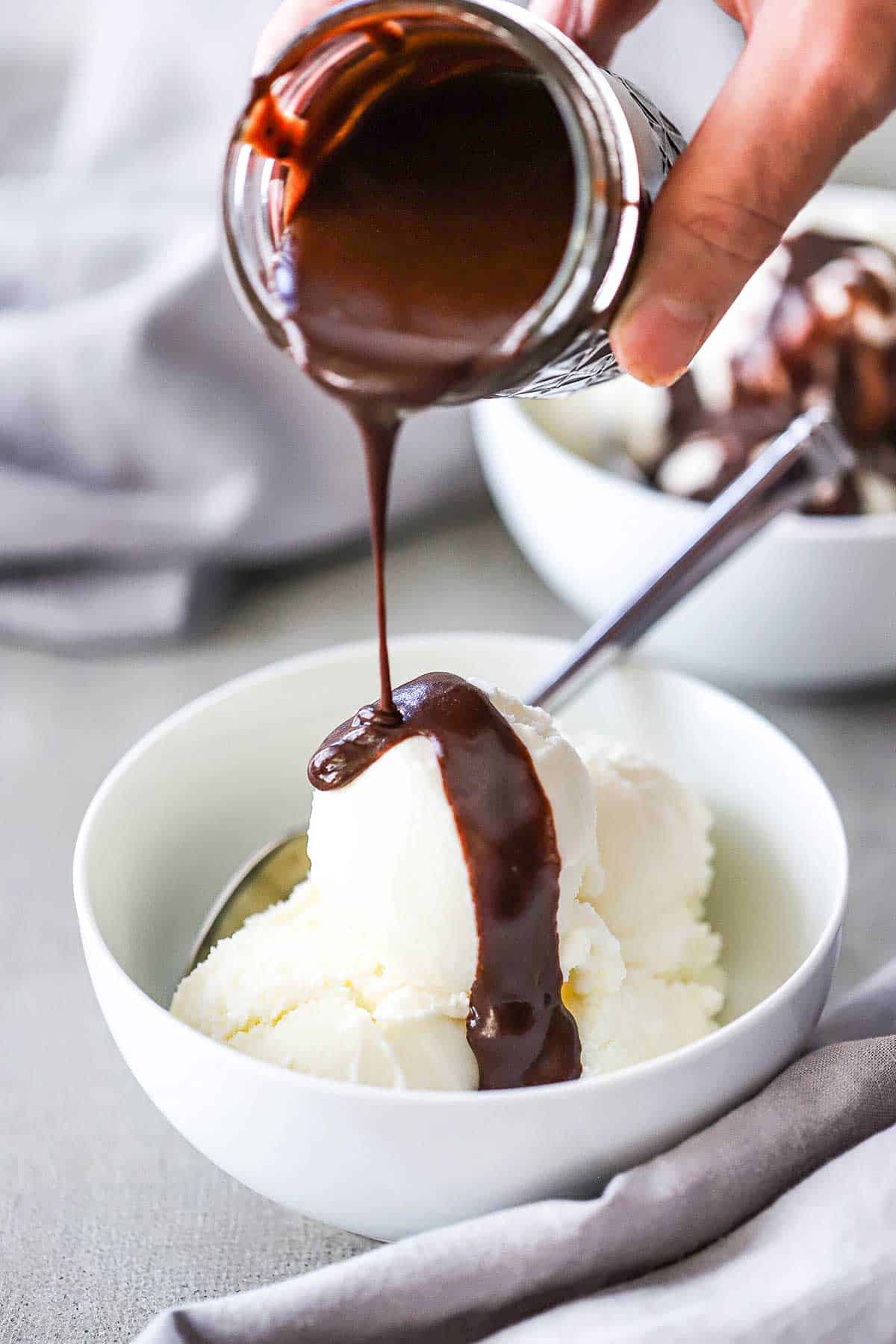 A person pouring chocolate sauce from a small glass jar over several scoops of homemade vanilla ice cream in a white ice cream bowl.