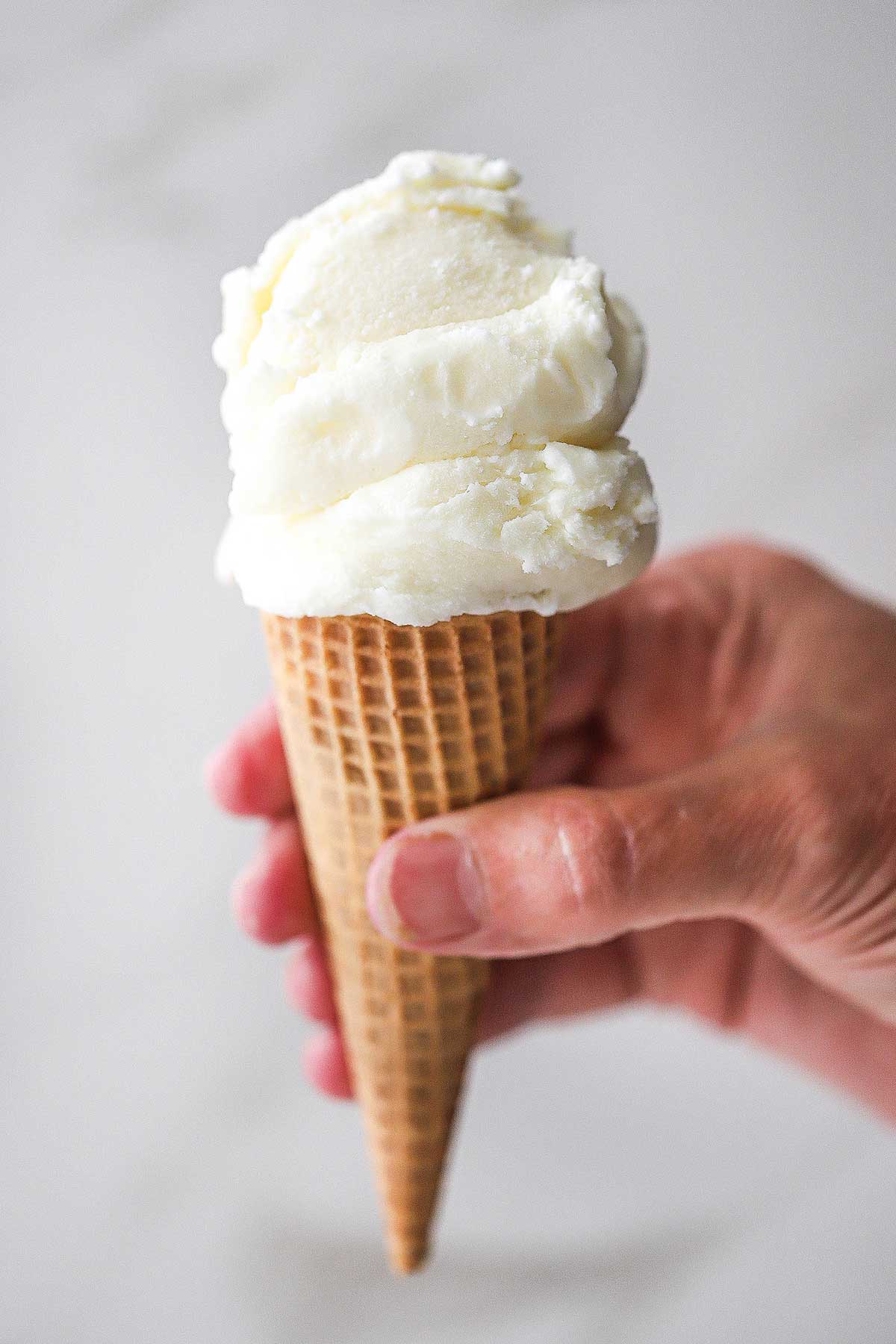 A person holding a sugar cone holding two scoops of vanilla ice cream on the top of the cone.