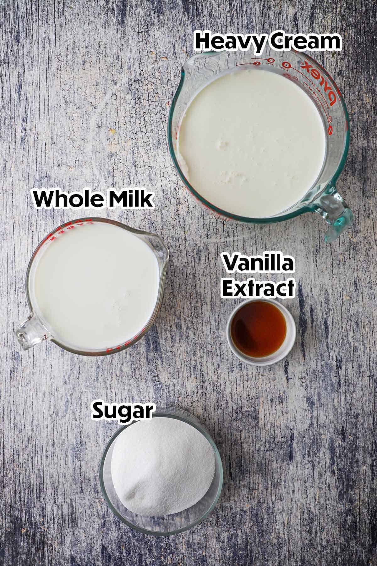 An arrangement of ingredients for homemade vanilla ice cream on a grey wooden background including heavy cream, whole milk sugar, and vanilla extrac.
