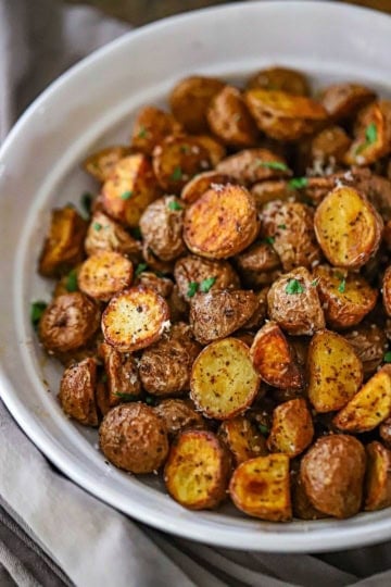 An close-up view of a white shallow bowl filled with a mound of Crispy Air-Fryer Baby Potatoes that have been garnished with grated Parmesan cheese, sea salt, and chopped parsley.
