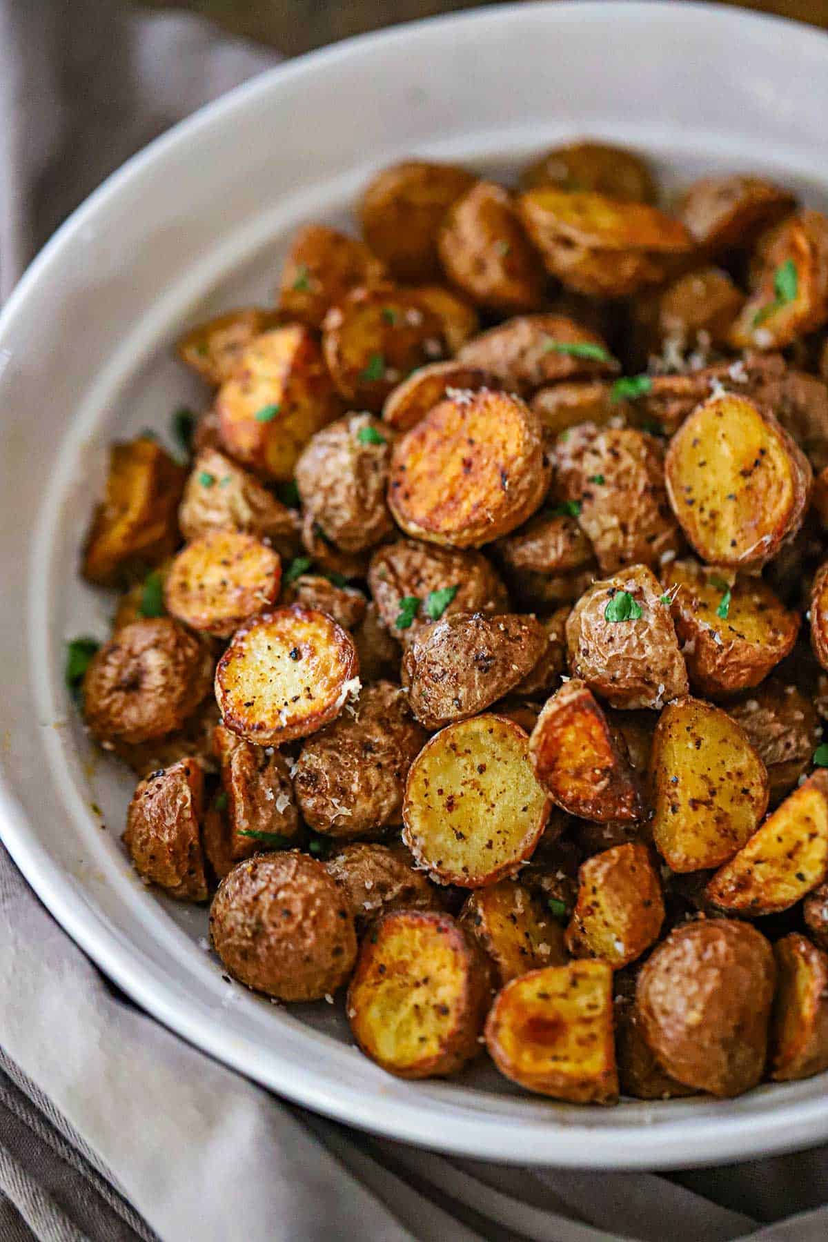 An overhead view of a white shallow bowl filled with a mound of Crispy Air-Fryer Baby Potatoes that have been garnished with grated Parmesan cheese, sea salt, and chopped parsley.