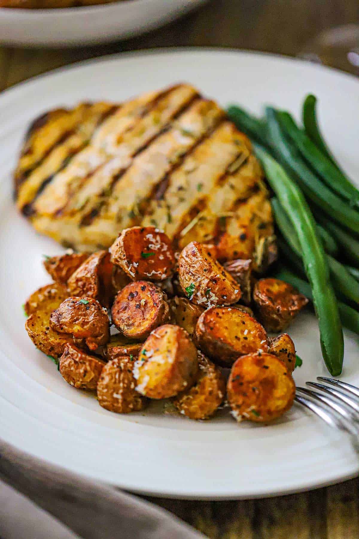 A straight-on view of a white dinner plate filled with a mound of Crispy Air-Fryer Baby Potatoes next to steamed green beans and a grilled chicken breast.