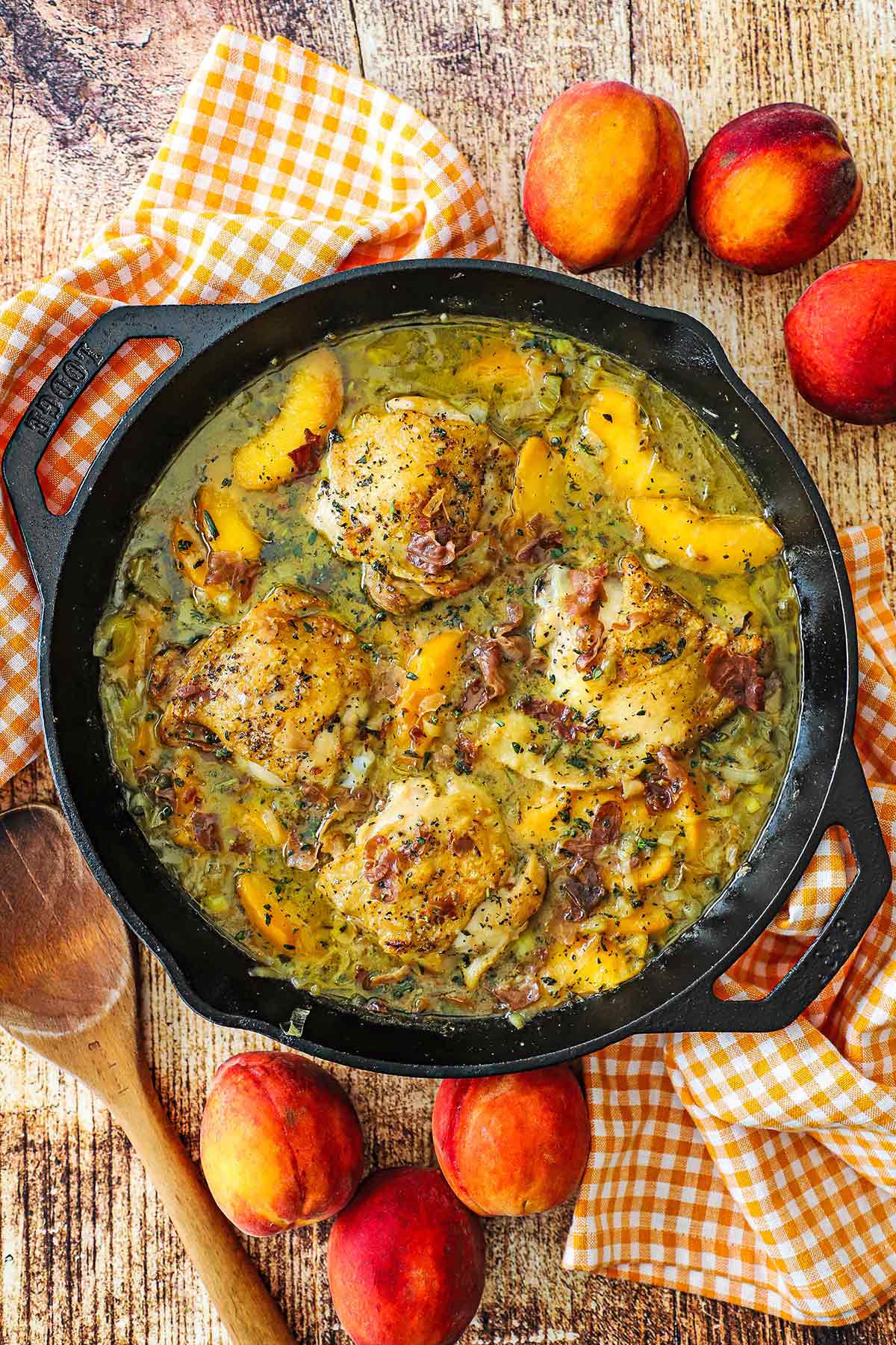 An overhead view of braised chicken and peaches in a large cast-iron skillet with fresh peaches placed around the skillet.