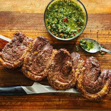 Four freshly cooked Brazilian Picanha steaks on a skewer resting on a cutting board with a bowl and spoon filled with homemade chimichurri sauce next to it.