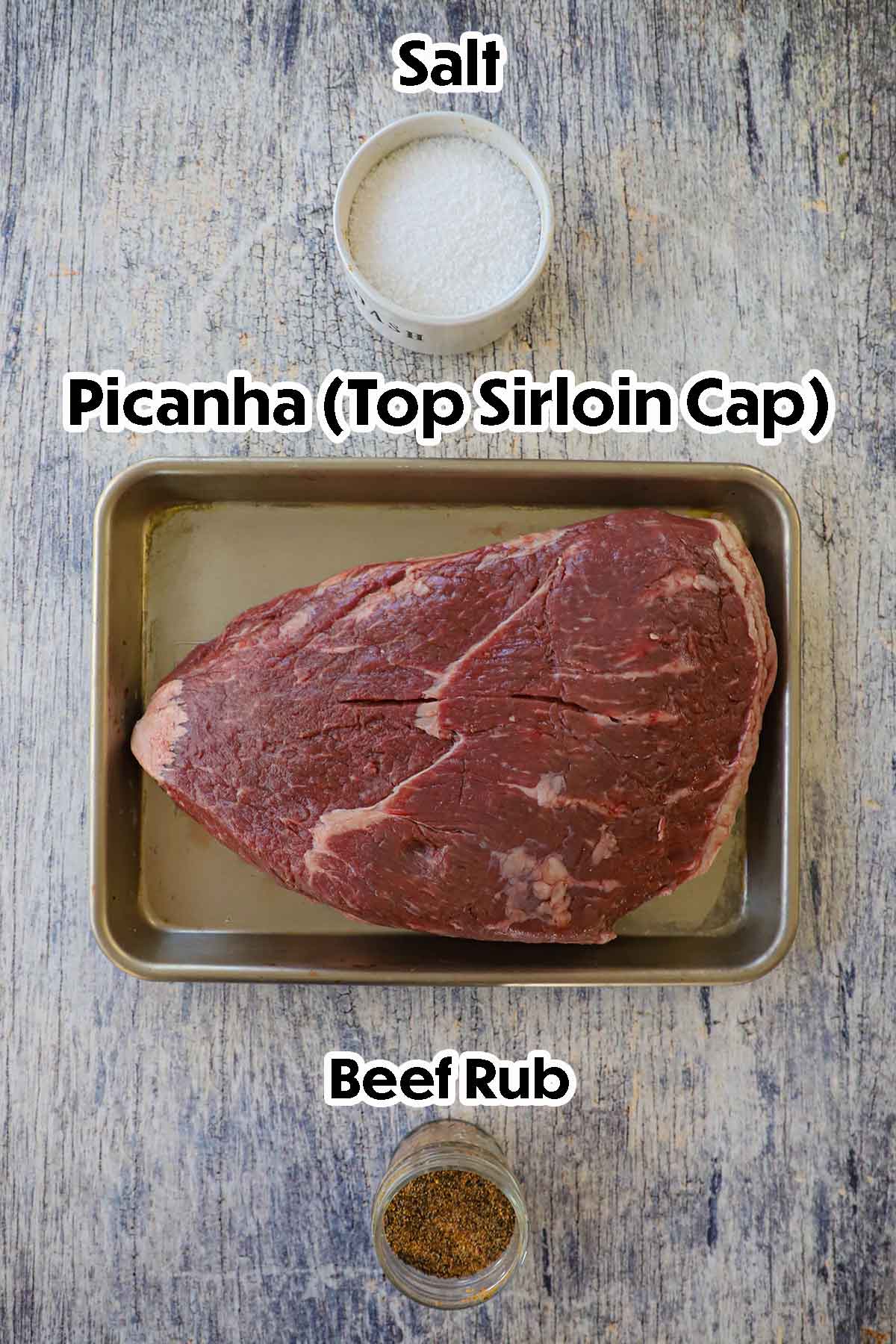 An arrangement of ingredients for Brazilian Picanha on a grey wooden background including an uncooked picanha steak resting in a metal tray, salt, and a jar filled beef rub.