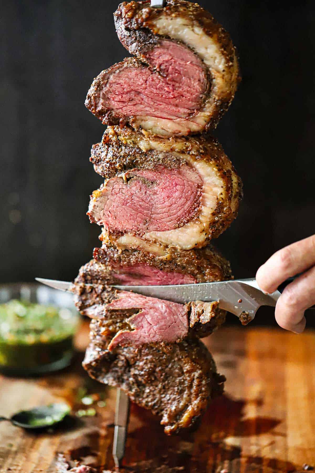 A person using a sharp carving knife to slice thin layers of picanha steak from a one of three folded steaks that are all on a skewer and freshly baked with a rotisserie.