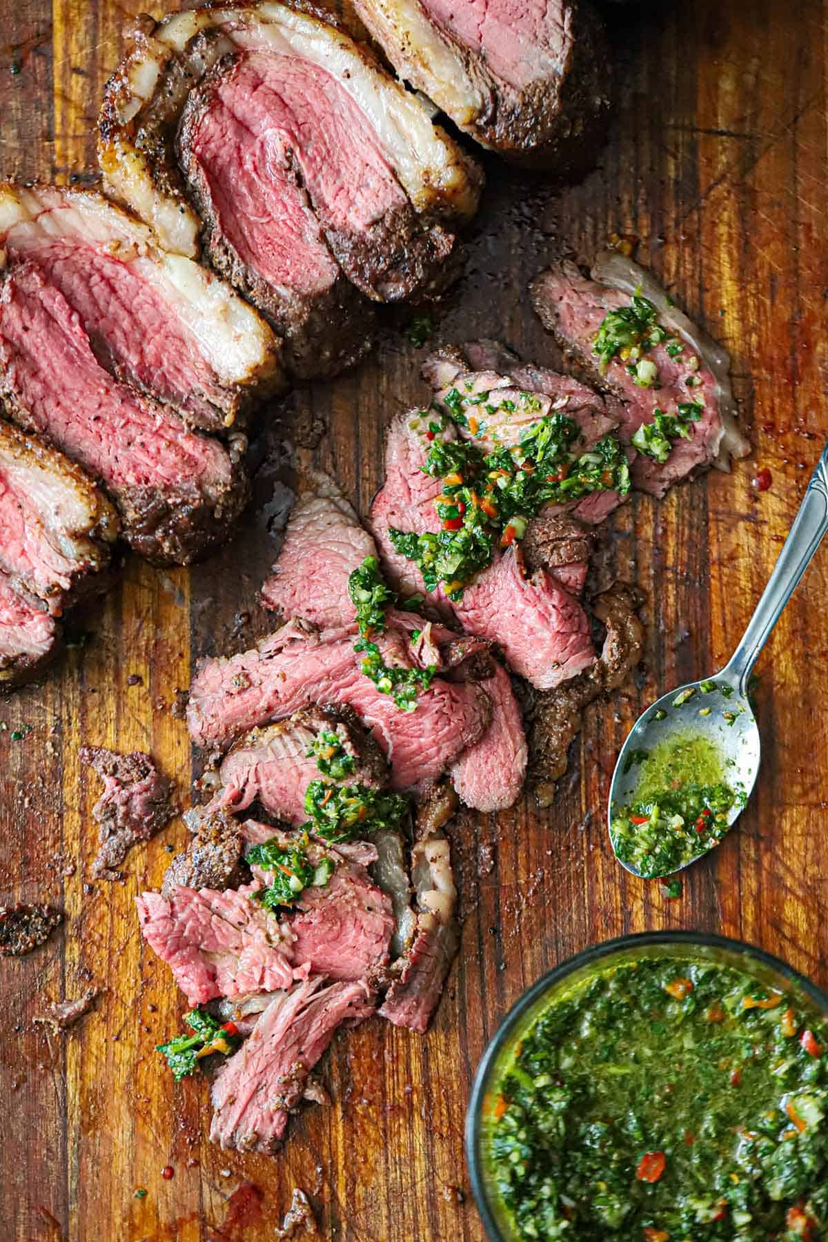 An overhead view of a row of thinly slice medium-rare picanha steak on a cutting board with blobs of homemade chimichurri sauce layered down the middle of the meat.