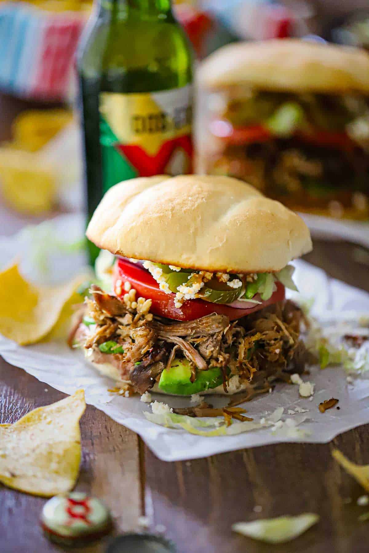 A straight-on view an overflowing carnitas torta packed with layers of refried beans, avocado, carnitas, tomatoes, jalapeños, Cotija cheese, all on a homemade telera bun.