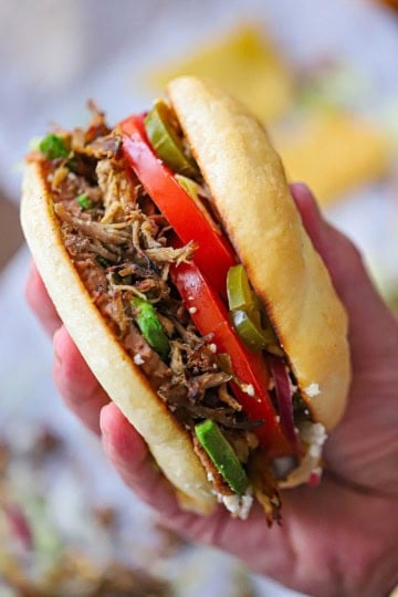 A person holding a homemade carnitas torta sandwich in the palm of his hand with the shredded pork filling and juicy vegetables clearly visible.