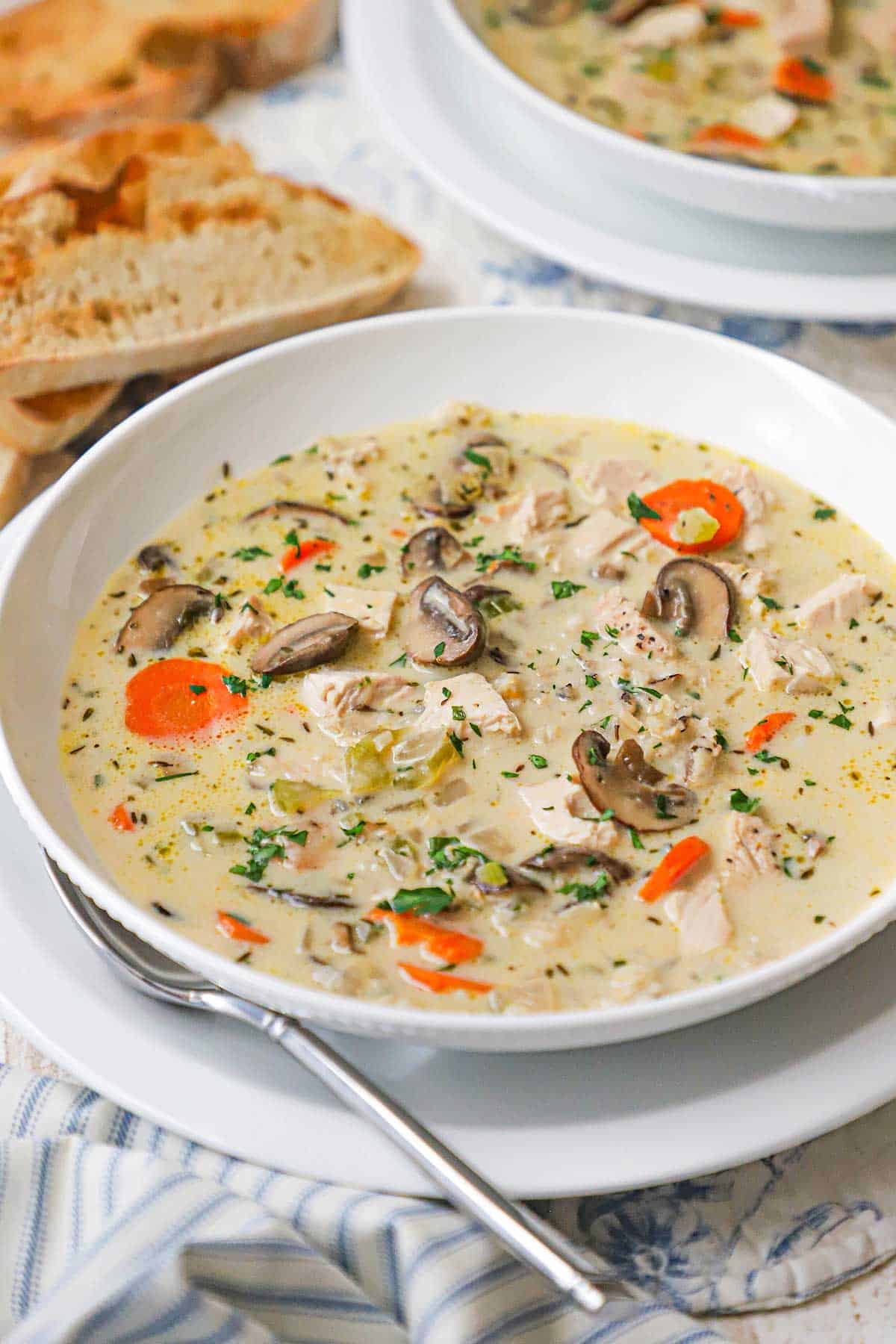A straight-on view of a shallow white bowl filled with a serving of Creamy Chicken and Wild Rice Soup with several sliced of toasted bread in the background.
