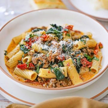 A close-up view of a shallow bowl filled with a serving of Tuscan Sausage Pasta loaded with spinach, sun-dried tomatoes, and crumbled sausage topped with grated Parmesan cheese.