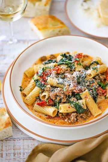 A straight-on view of a shallow bowl filled with a serving of Tuscan Sausage Pasta loaded with spinach, sun-dried tomatoes, and crumbled sausage topped with grated Parmesan cheese.