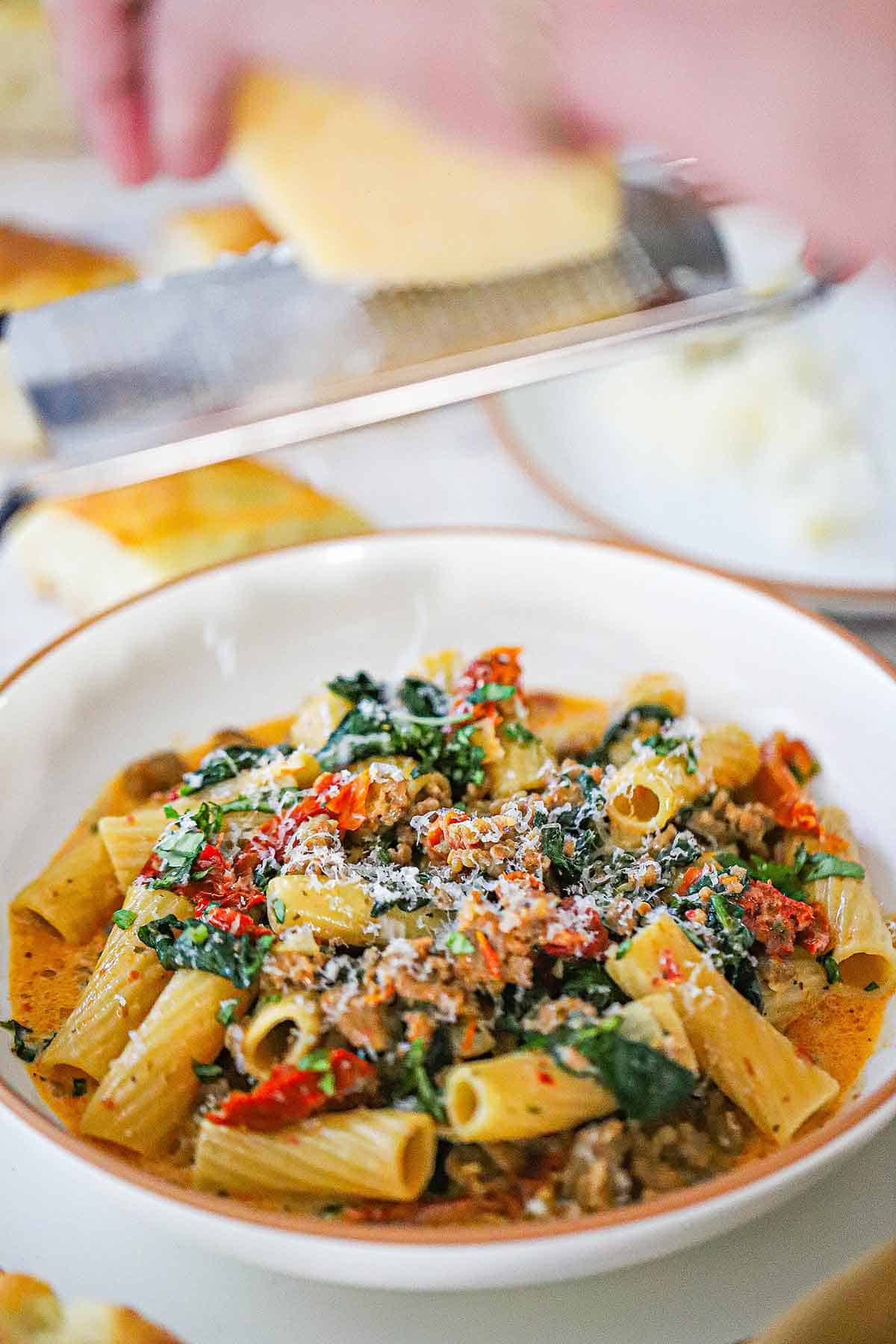 A person using a microplane to grate a block of Parmesan cheese over a shallow pasta bowl filled with a serving of Tuscan Sausage Pasta.