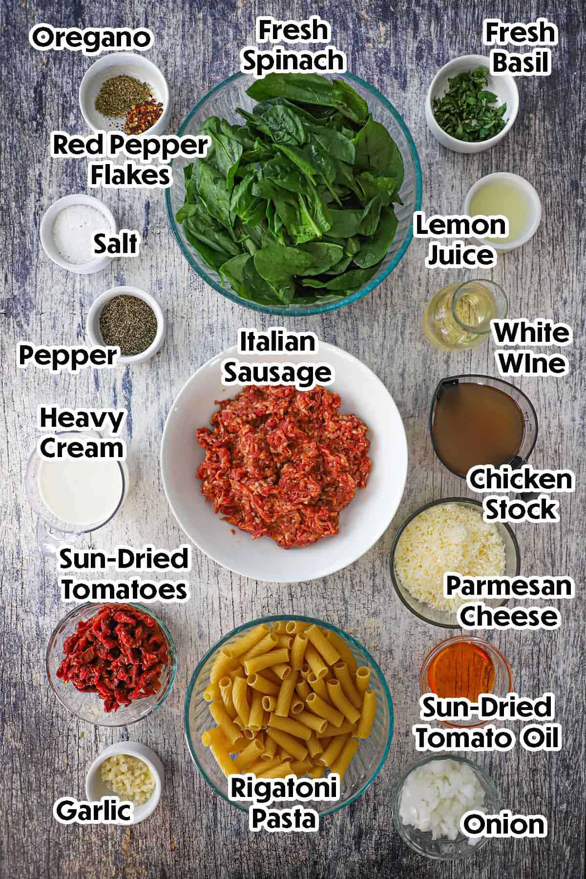 An arrangement of ingredients for Tuscan Sausage Pasta on a grey wooden background including Italian sausage, fresh spinach, pasta, cream, chicken stock, sun-dried tomatoes, garlic, cheese, white wine, herbs, and seasonings.