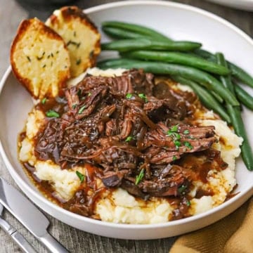 A straight-on view of a shallow dinner bowl filled with a serving of French onion pot roast resting on a bed of mashed potatoes next to steamed green beans and two pieces of toasted baguette slices.