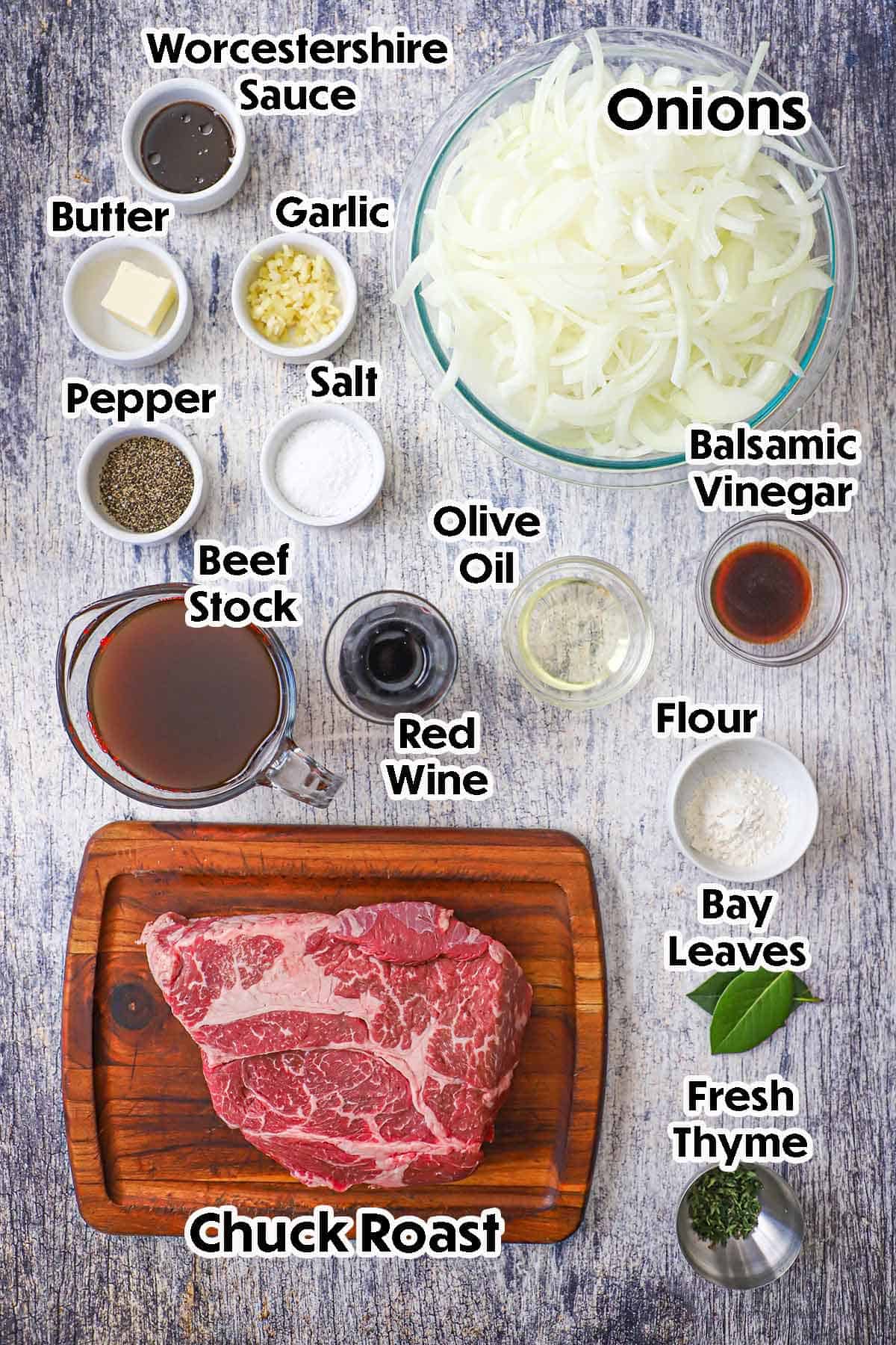 An arrangement of ingredients for French Onion Pot Roast on a grey wooden background including a chuck roast, onions, beef stock, garlic, butter, balsamic vinegar, flour, red wine, and seasonings.