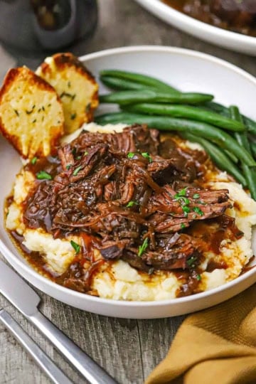 A straight-on view of a shallow dinner bowl filled with a serving of French onion pot roast resting on a bed of mashed potatoes next to steamed green beans and two pieces of toasted baguette slices.