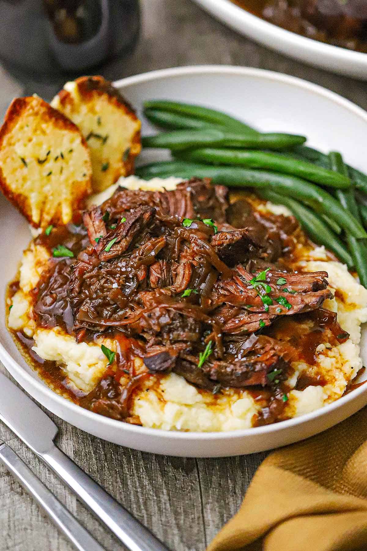 A straight-on view of a shallow dinner bowl filled with a serving of French onion pot roast resting on a bed of mashed potatoes next to steamed green beans and two pieces of toasted baguette slices.