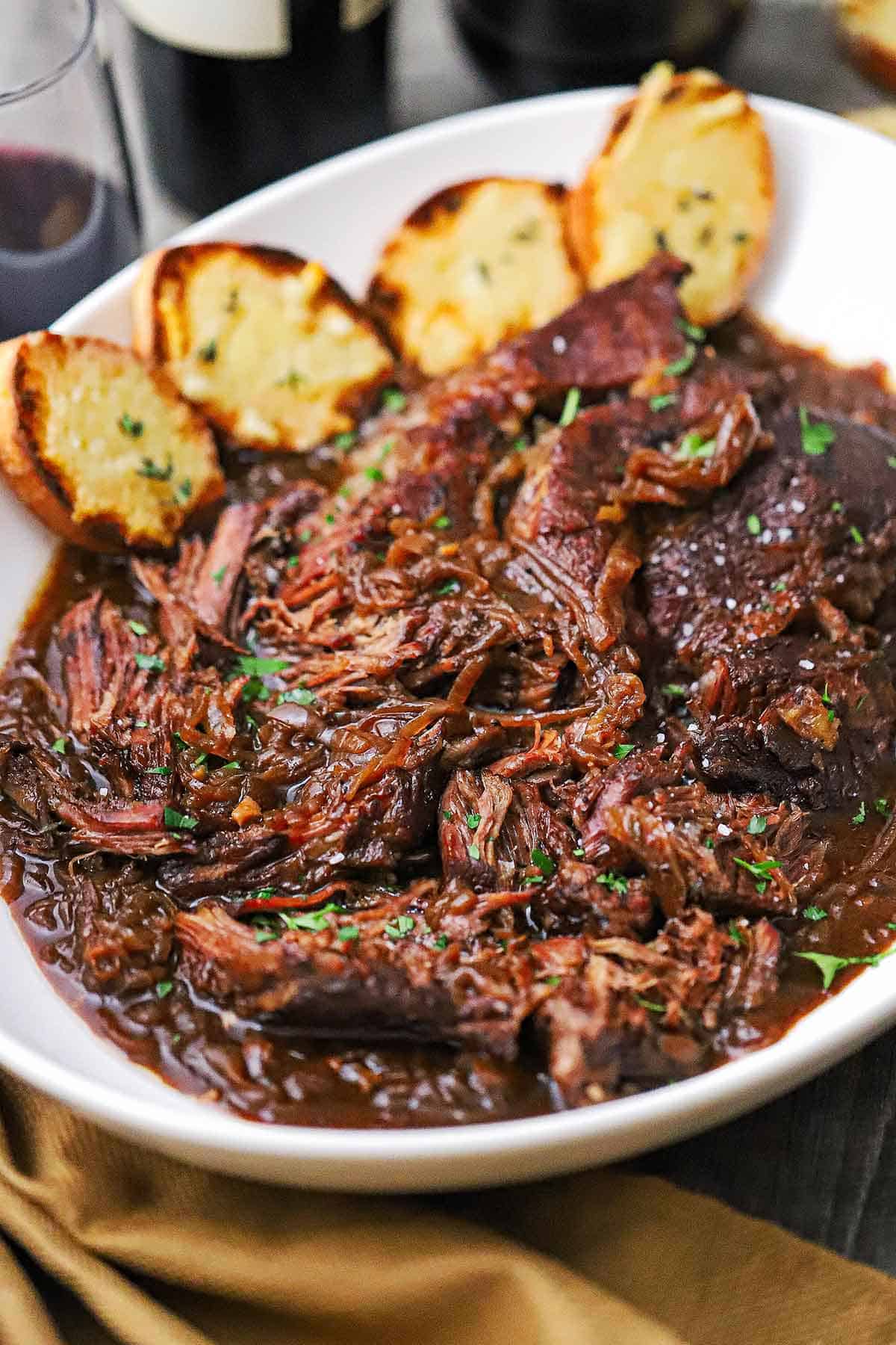 A large oval platter filled with a shredded French onion pot roast garnished with chopped Italian parsley with four slices of toasted bread resting on the side of the dish.