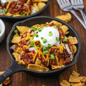 A close-up view of a small cast-iron skillet filled with an individual serving of Classic Frito Pie topped with Beef Chili, shredded cheese, sour cream, and chopped jalapeños.