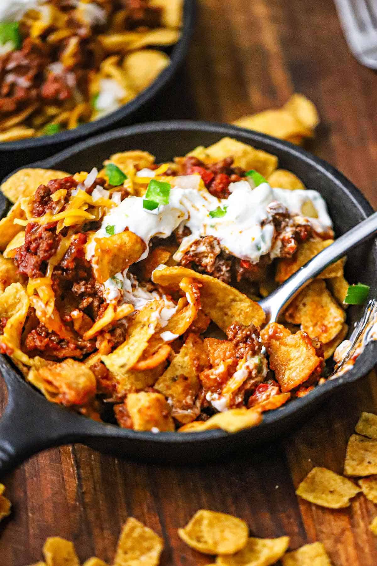 A staight-on view of a small cast-iron skillet filled with a partially eaten serving of Classic Frito Pie topped with chopped jalapeños and sour cream.